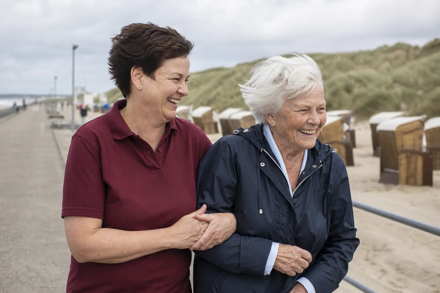 Seniorin und Betreuerin spazieren lachend an einer Strandpromenade