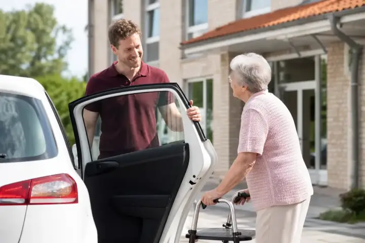 Fahrdienst: Betreuer hält einer Seniorin mit Rollator die Autotür auf